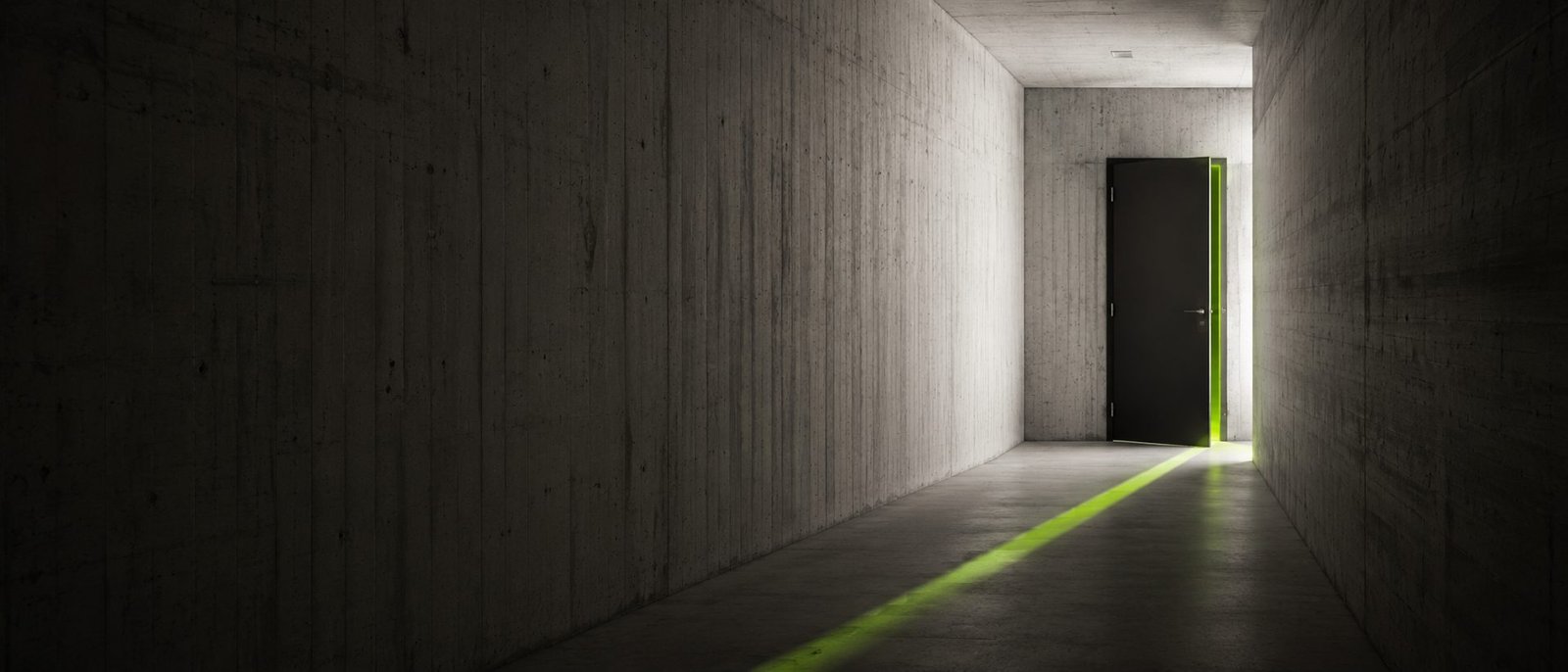 Corredor de concreto em penumbra com uma porta entreaberta ao fundo, de onde escapa uma faixa de luz verde sobre o piso